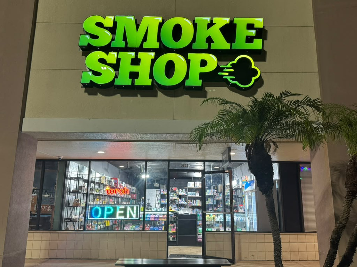 iSmokee Orlando storefront at night — the lit green SMOKE SHOP sign above the entrance, OPEN neon in the window and the stocked shelves visible inside